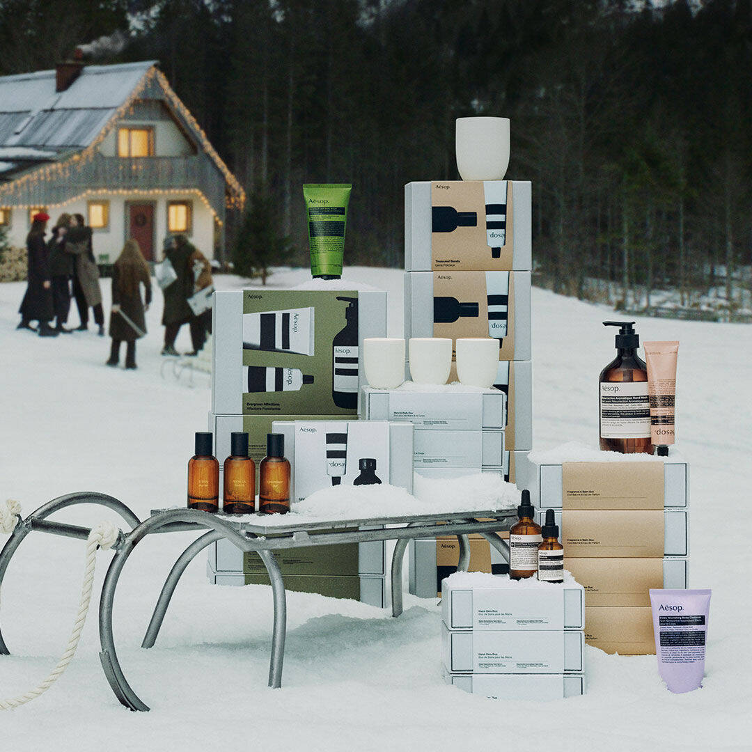 Aesop gift sets and products arranged on and around a sled in the snow, with a festively lit chalet and forest in the background.