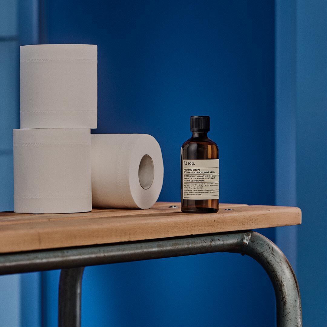 Aesop Post-Poo Drops bottle beside stacked toilet rolls on a wooden shelf against a blue wall.