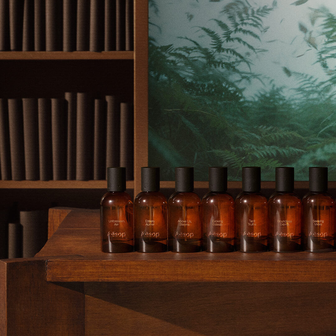 Row of Aesop amber fragrance bottles with black caps lined up on a dark wooden shelf, with cylindrical wooden elements on shelves and a green fern panel in the background.