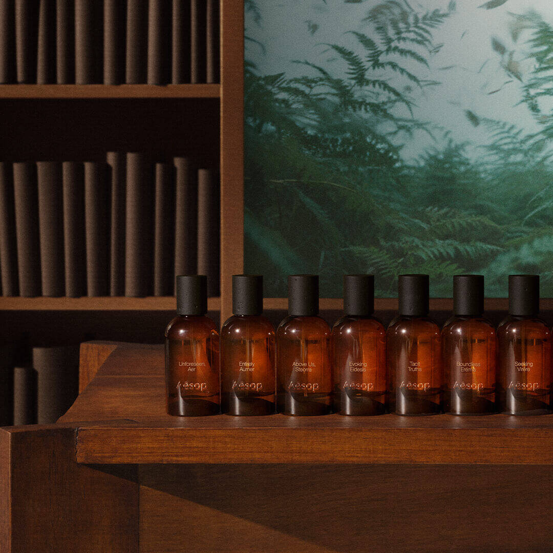 Row of Aesop amber fragrance bottles with black caps lined up on a dark wooden shelf, with cylindrical wooden elements on shelves and a green fern panel in the background.