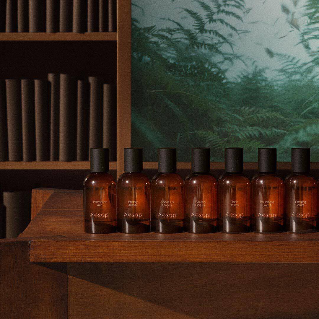 Row of Aesop amber fragrance bottles with black caps lined up on a dark wooden shelf, with cylindrical wooden elements on shelves and a green fern panel in the background.