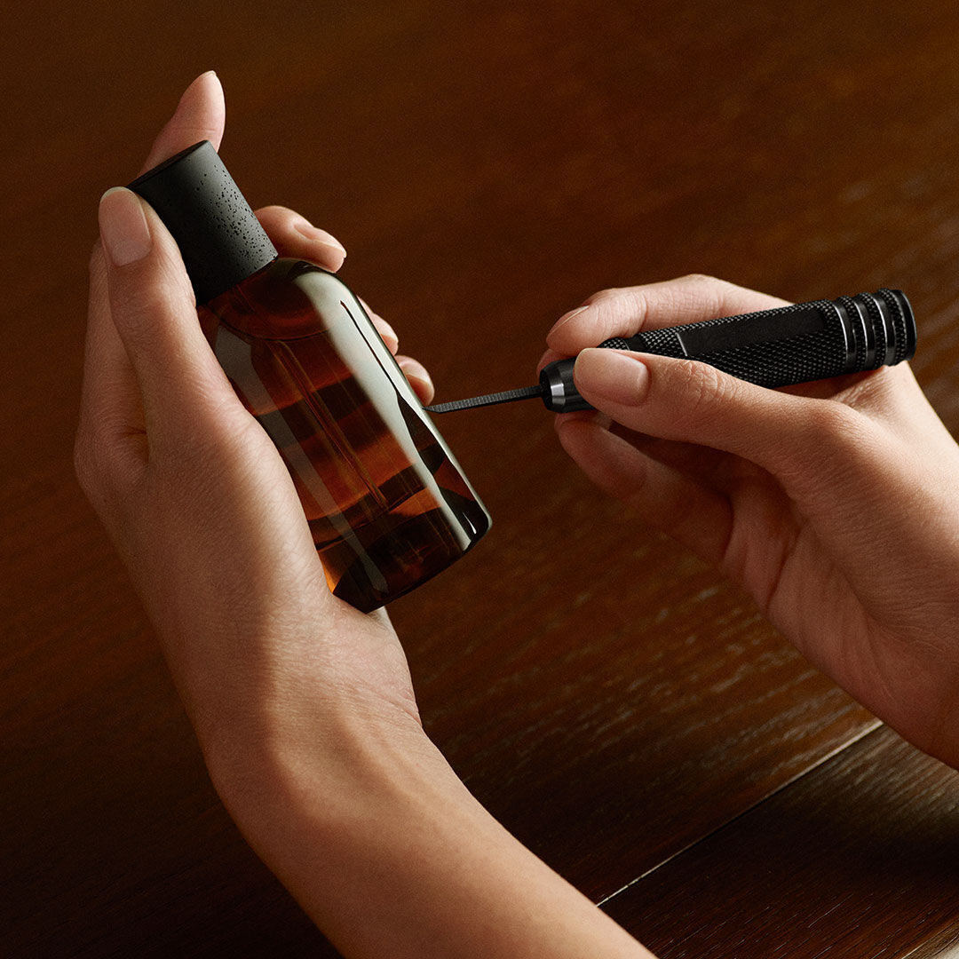 Hands hold an amber glass bottle and a black engraving tool, its tip touching the bottle above a wooden surface.