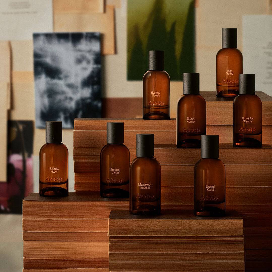 Collection of Aesop amber fragrance bottles with black caps arranged on tiered wooden stepped display platforms against a collage backdrop of prints and paintings.