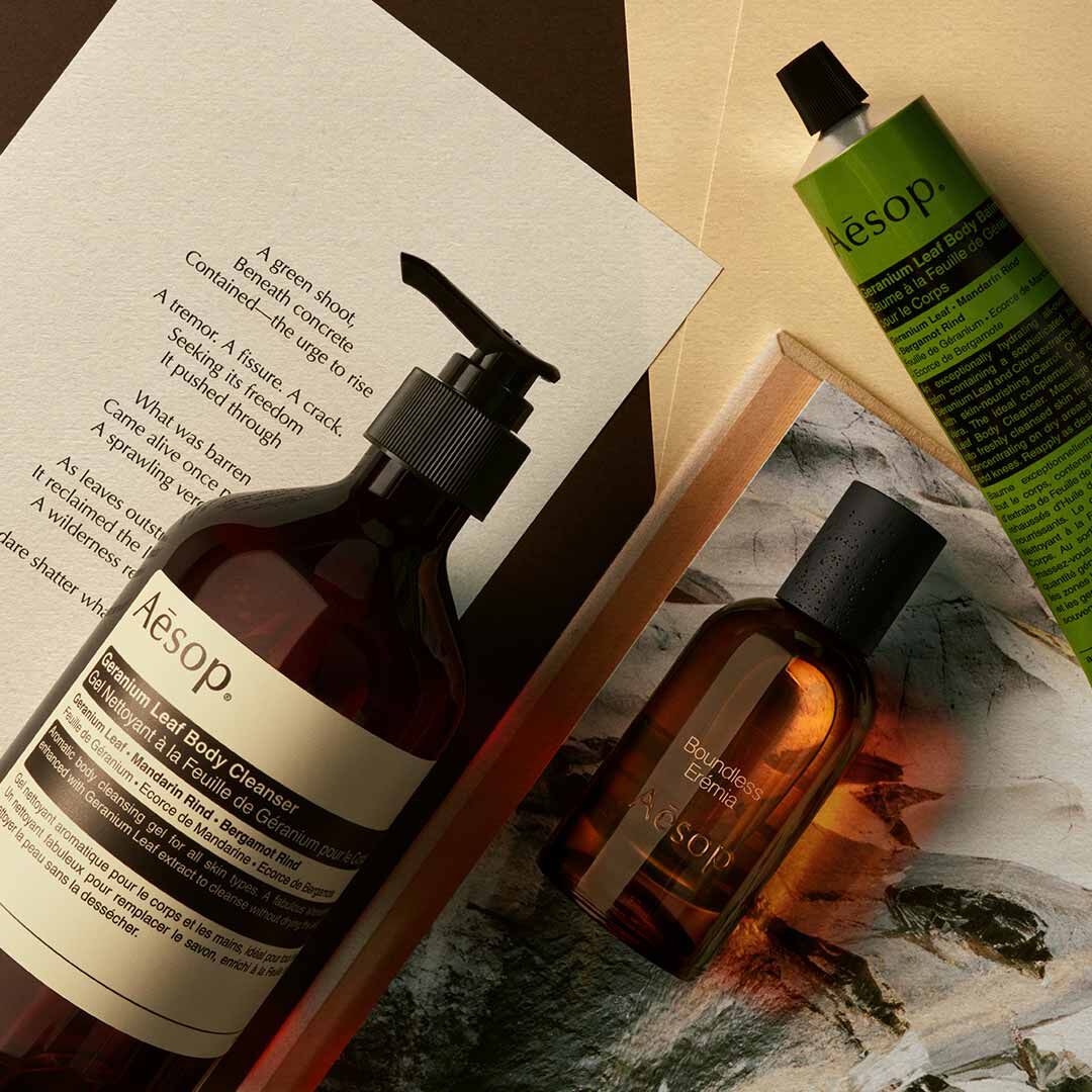 Aesop Boundless, Eremia fragrance bottle, large amber pump bottle and cosmetic tube on open books and printed pages.