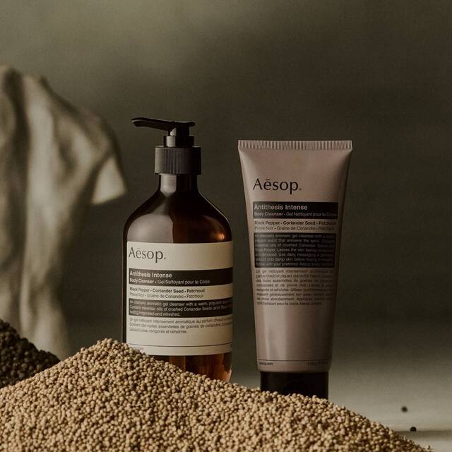 Aesop Antithesis Intense Body Cleanser pump bottle and tube beside piles of black peppercorns and coriander seeds.
