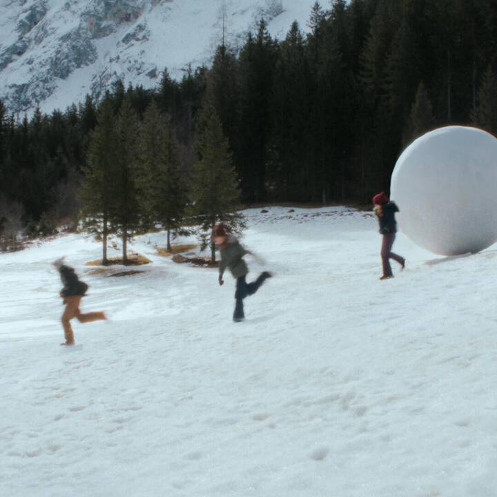 People running across snow towards a large white sphere, with evergreen trees and snow-covered mountains behind them.