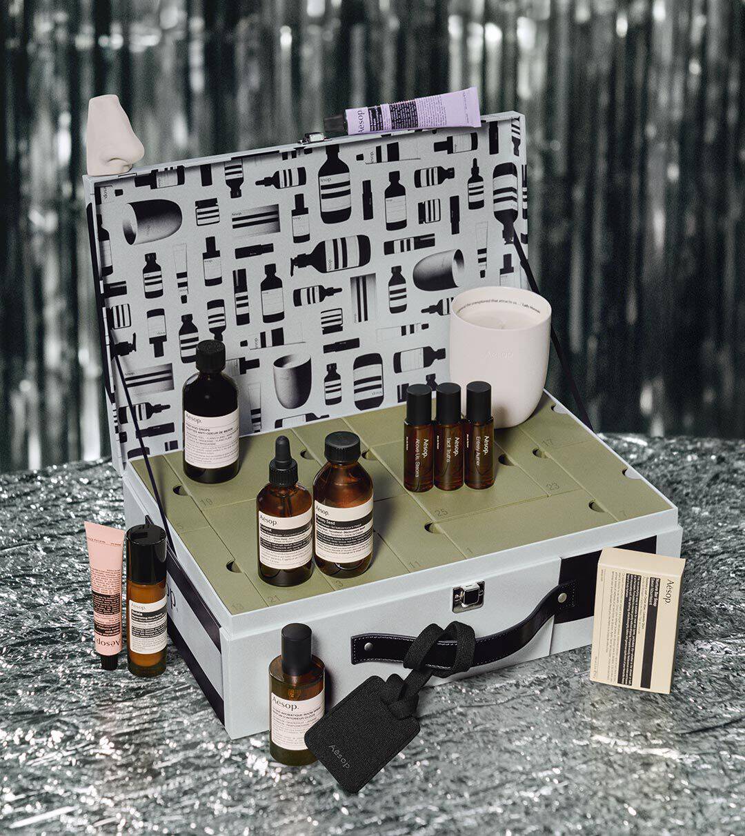 Aesop bottles, tubes and a white jar arranged in and around an open silver case with a patterned lid, against silver tinsel.