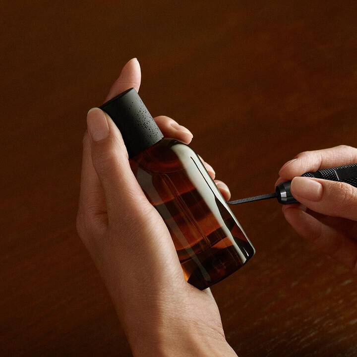 A close-up of hands engraving a dark amber Aesop fragrance bottle with a handheld tool against a warm wooden background.