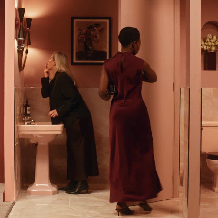 Two people in a softly lit pink-tiled bathroom with a toilet and sink.