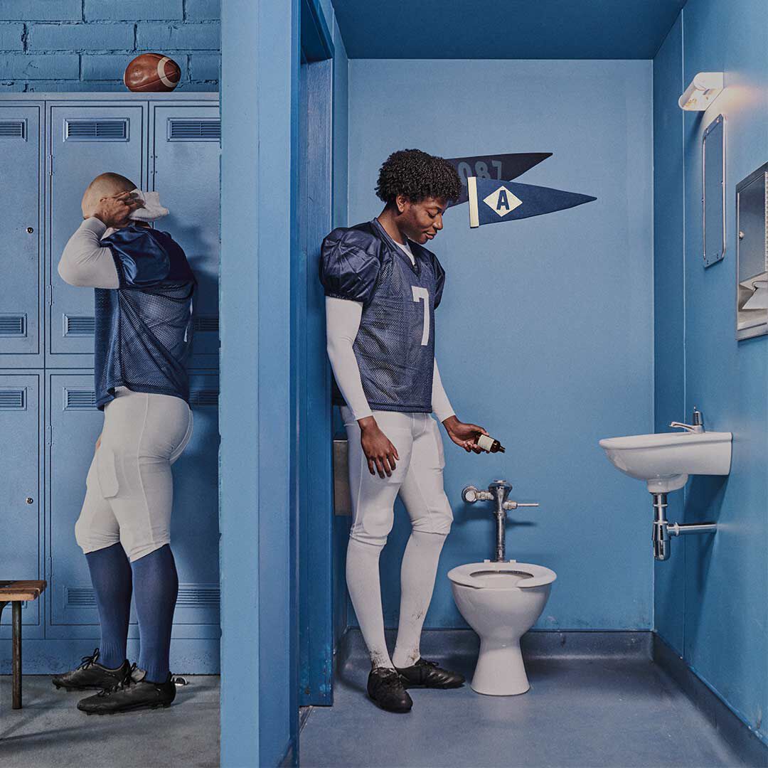 American football players in a blue locker room beside a toilet with Aesop Post-Poo Drops.