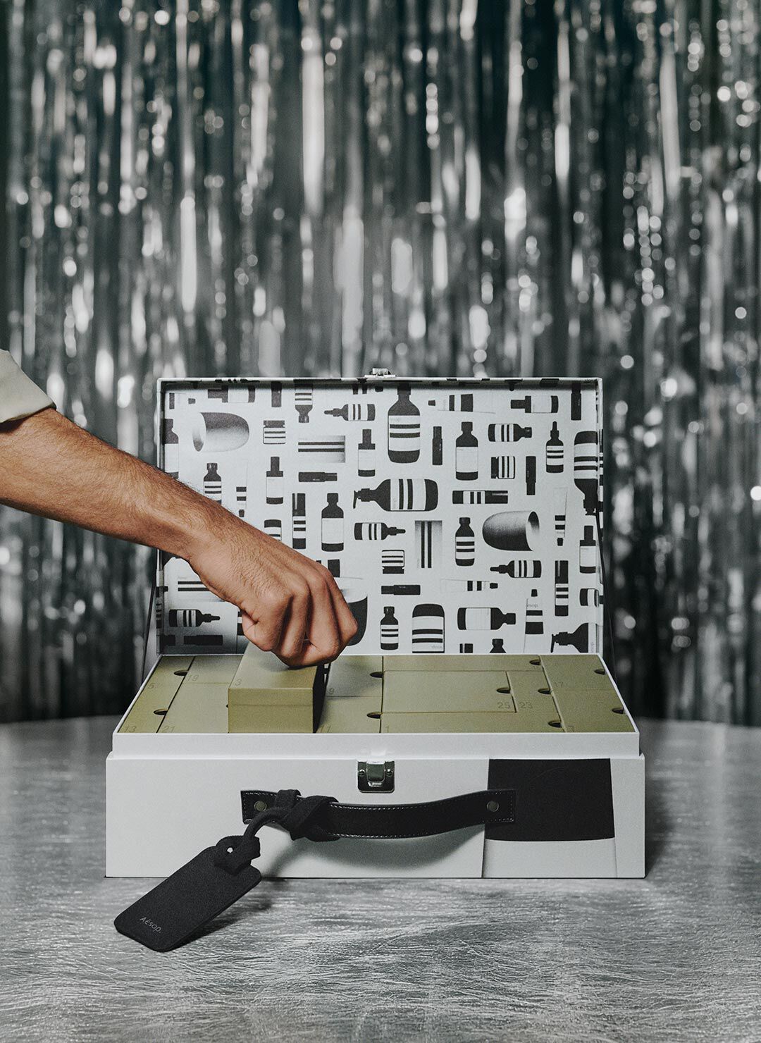 Hand lifting a green compartment lid inside an open silver case with a black-and-white patterned interior, on a table against silver tinsel.