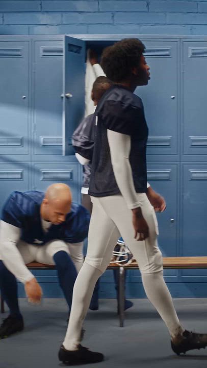American football players in a blue locker room, one bending beside a bench.