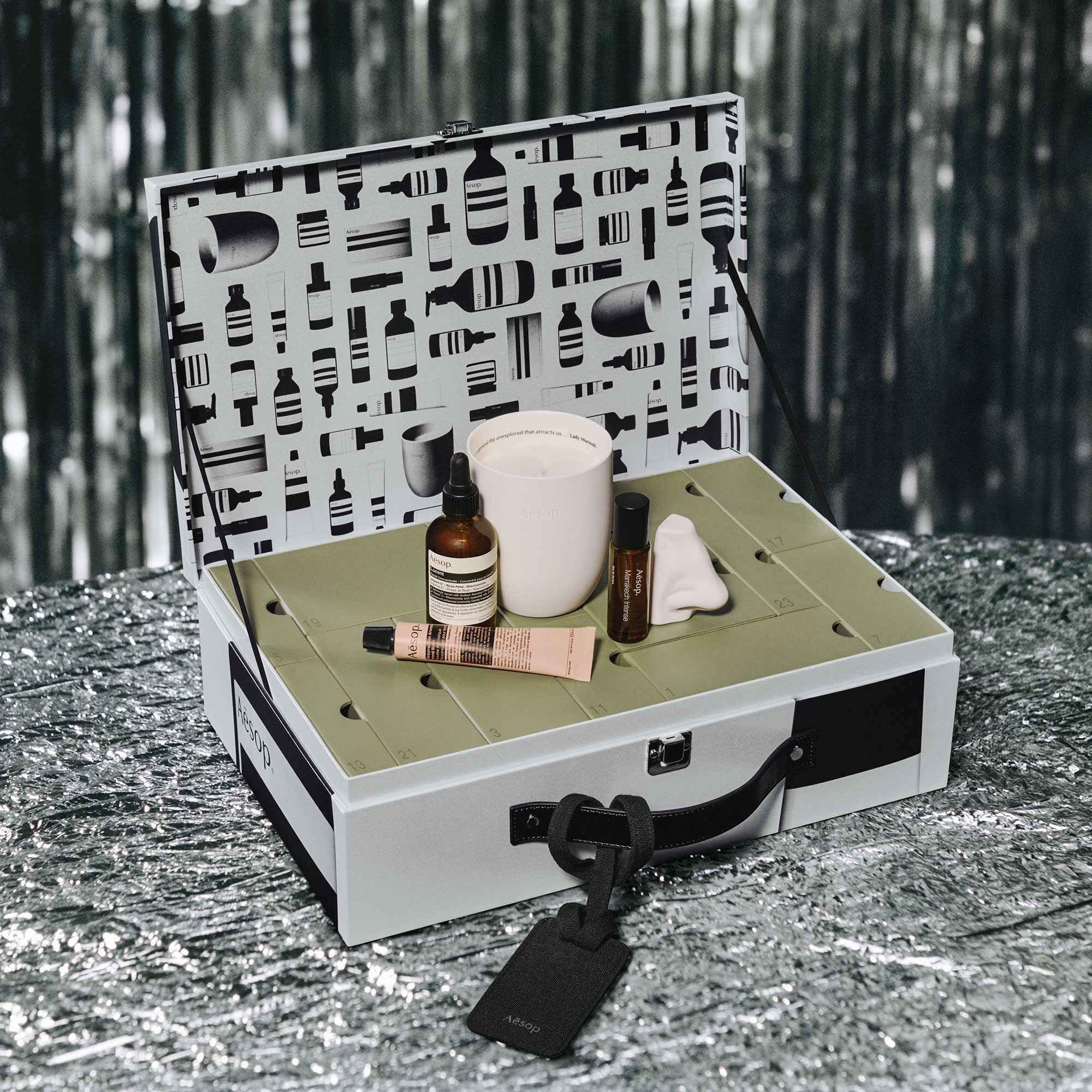 An amber dropper bottle, fragrance vial, pink tube, white candle and nose-shaped object rest on numbered boxes inside an open silver Aesop case.
