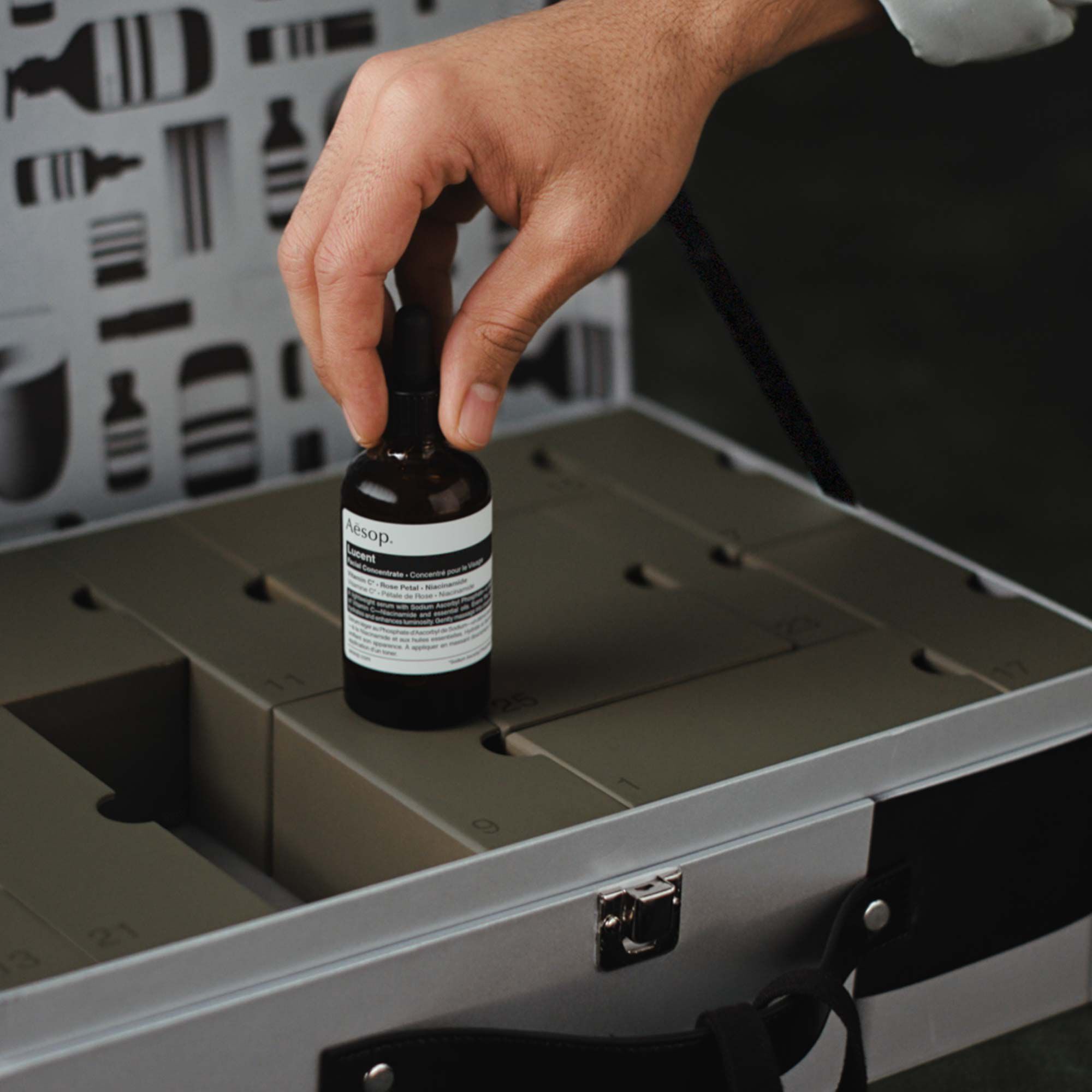 A hand holds an amber Aesop Lucent dropper bottle above olive-green numbered boxes inside an open silver case with a patterned lid.