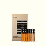 Six amber fragrance spray vials with black tops lined up before a gold Aesop Sharing Stories box on white.