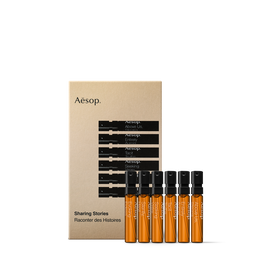 Six amber fragrance spray vials with black tops lined up before a gold Aesop Sharing Stories box on white.