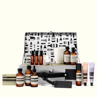 An open silver Aesop case with a patterned lid displays amber bottles, pastel tubes, boxed soap, a white candle and a nose-shaped object against white.