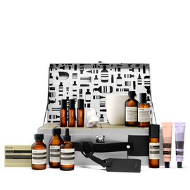 An open silver Aesop case with a patterned lid displays amber bottles, pastel tubes, boxed soap, a white candle and a nose-shaped object against white.