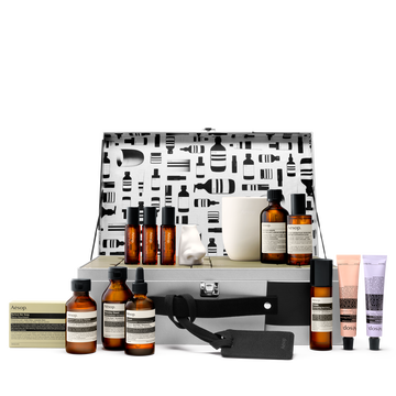 An open silver Aesop case with a patterned lid displays amber bottles, pastel tubes, boxed soap, a white candle and a nose-shaped object against white.