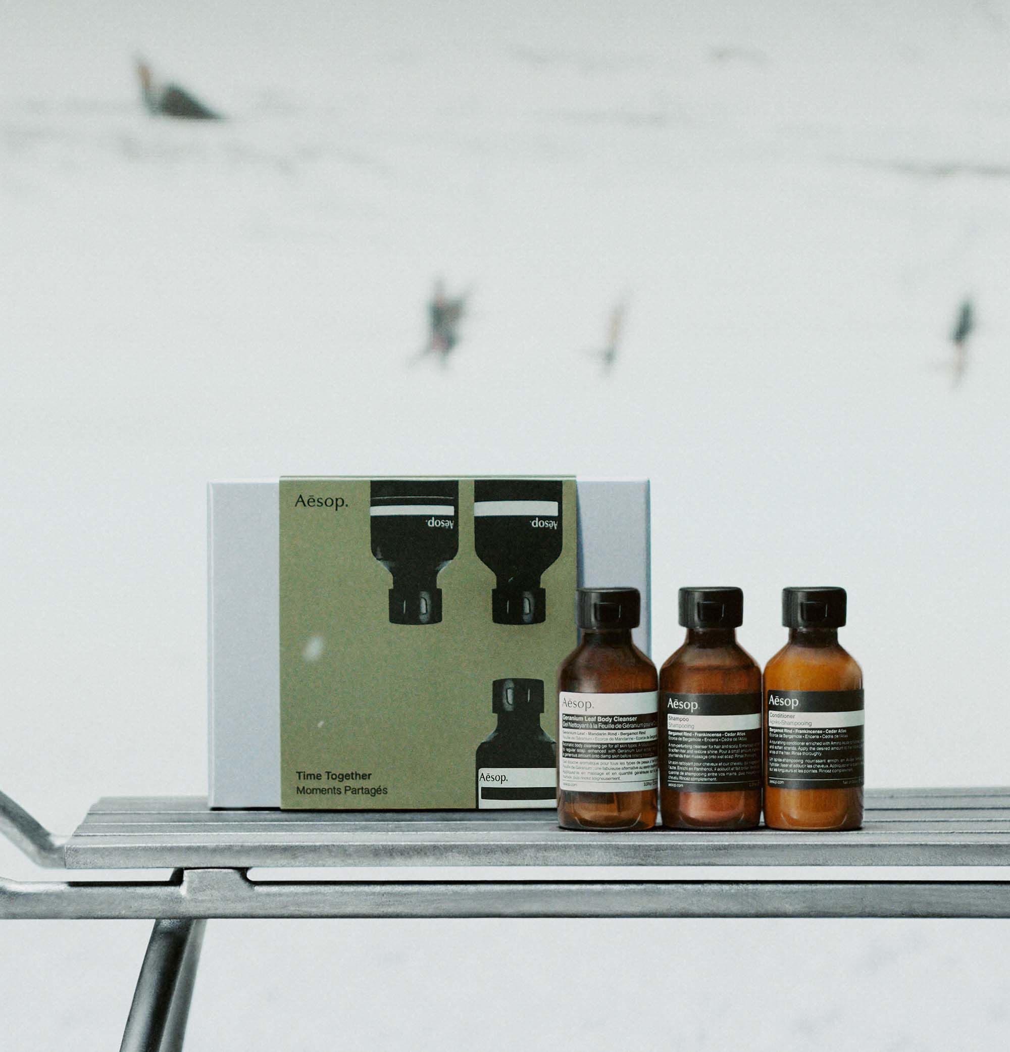 Three amber Aesop bottles with black caps stand beside a green and silver box on a metal sledge in snow.