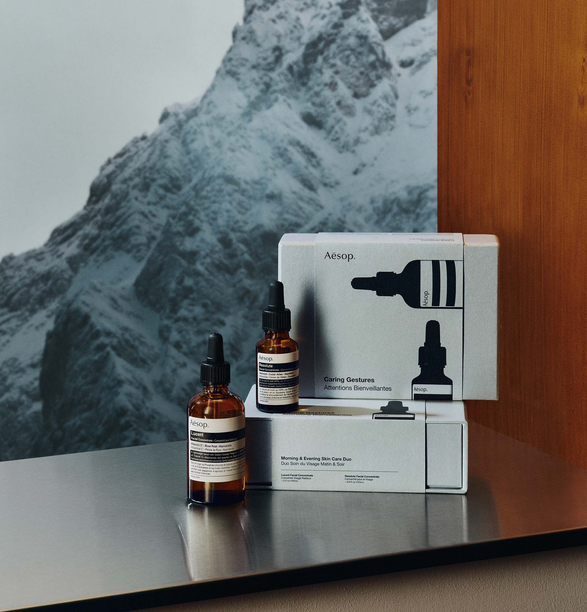 Two amber Aesop dropper bottles arranged at different heights beside stacked silver boxes, with snowy mountains beyond a reflective ledge.