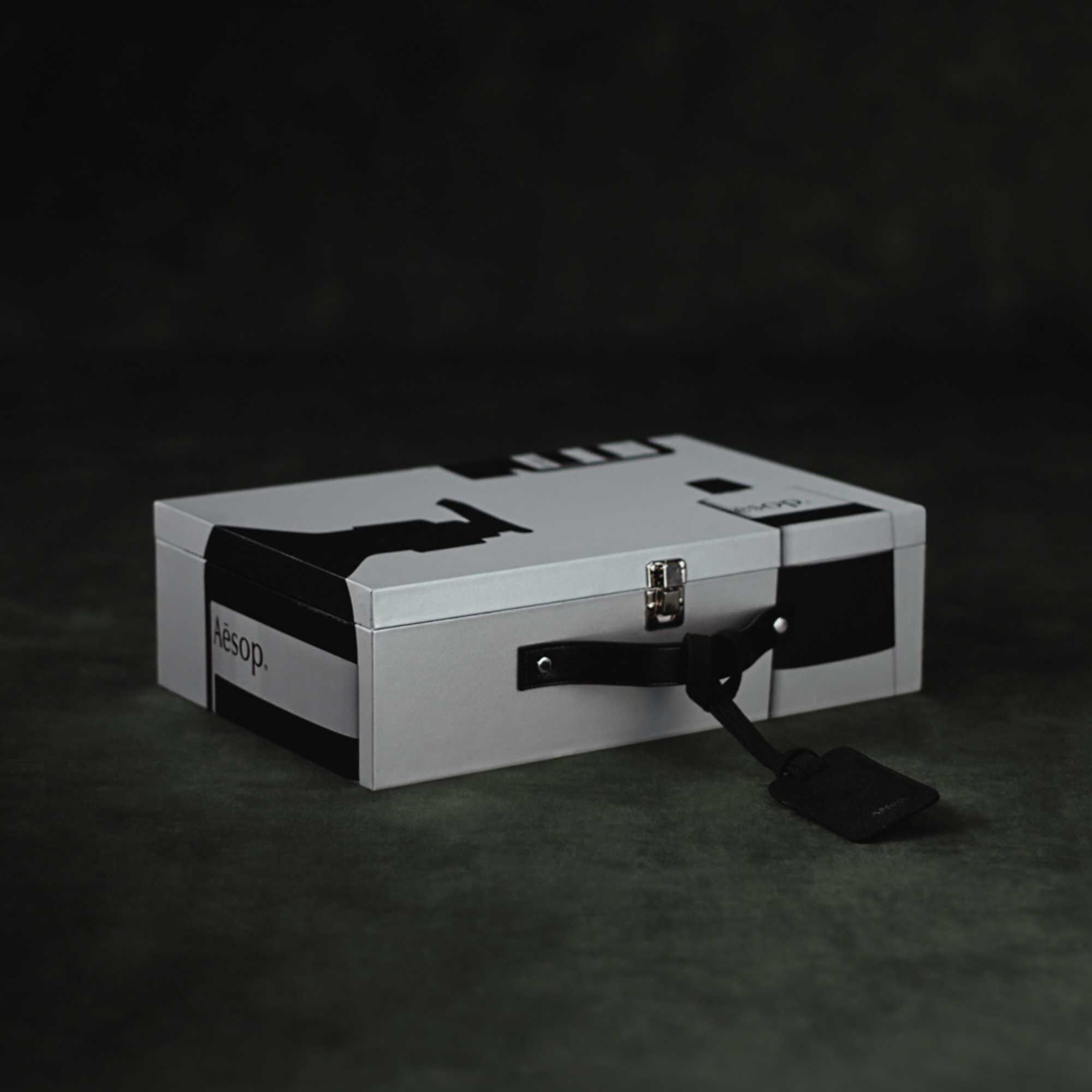 A closed silver Aesop case with black product illustrations, a black handle and luggage tag rests on a dark surface.
