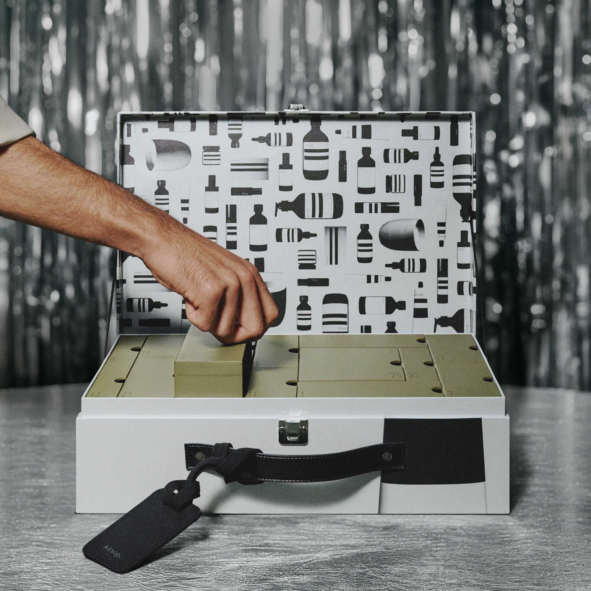 A hand lifts an olive-green numbered box from an open silver Aesop case with a patterned lid, against silver streamers.