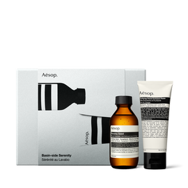 An amber Aesop bottle and cream-coloured tube with black caps stand in front of an illustrated silver-grey gift box.