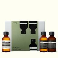 Three amber Aesop bottles with black caps stand around an olive-green and silver-grey box, one left and two right.
