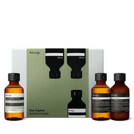 Three amber Aesop bottles with black caps stand around an olive-green and silver-grey box, one left and two right.