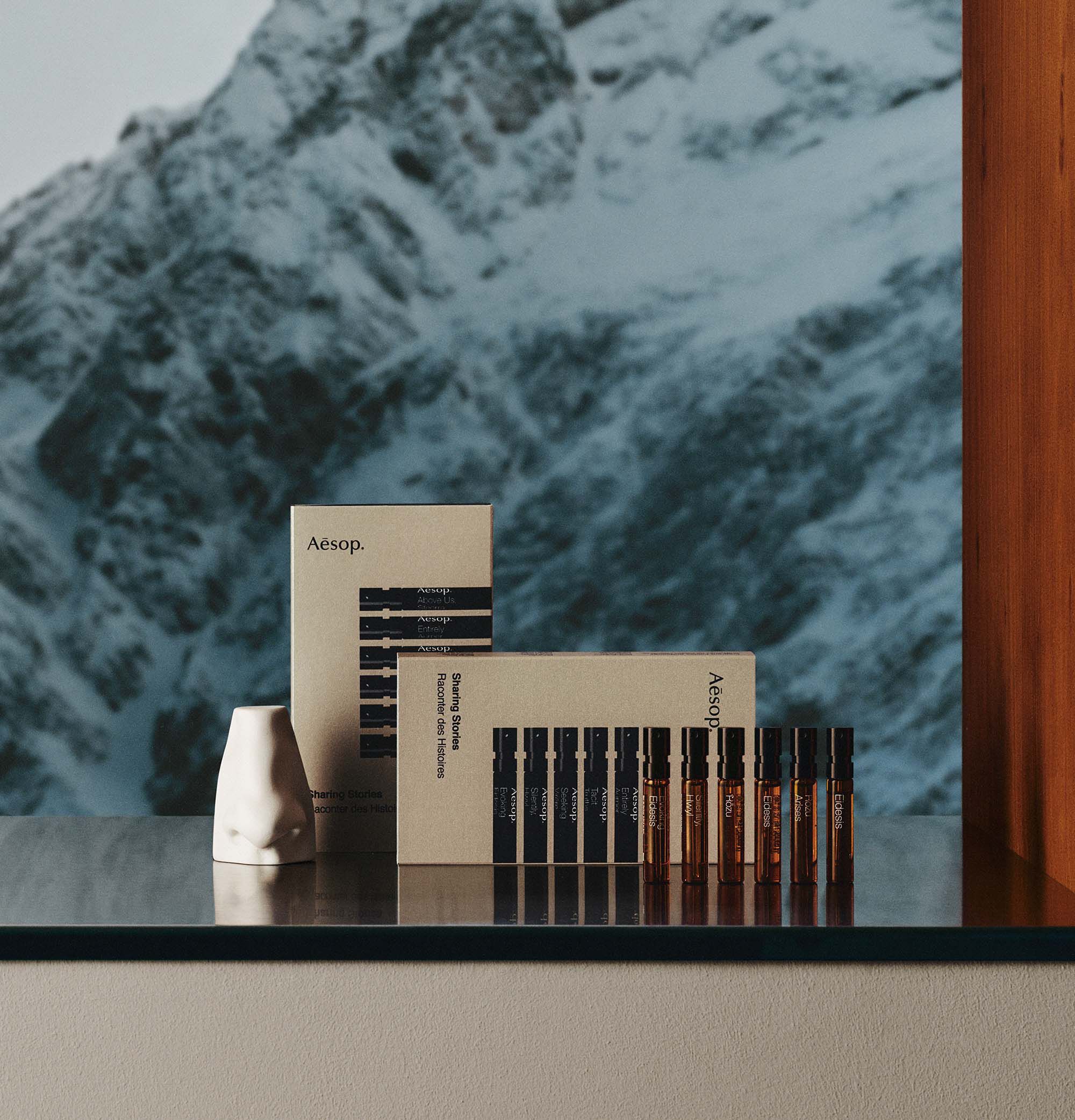 Six amber fragrance vials beside two gold Aesop boxes and a small white sculptural object, with snowy mountains behind.