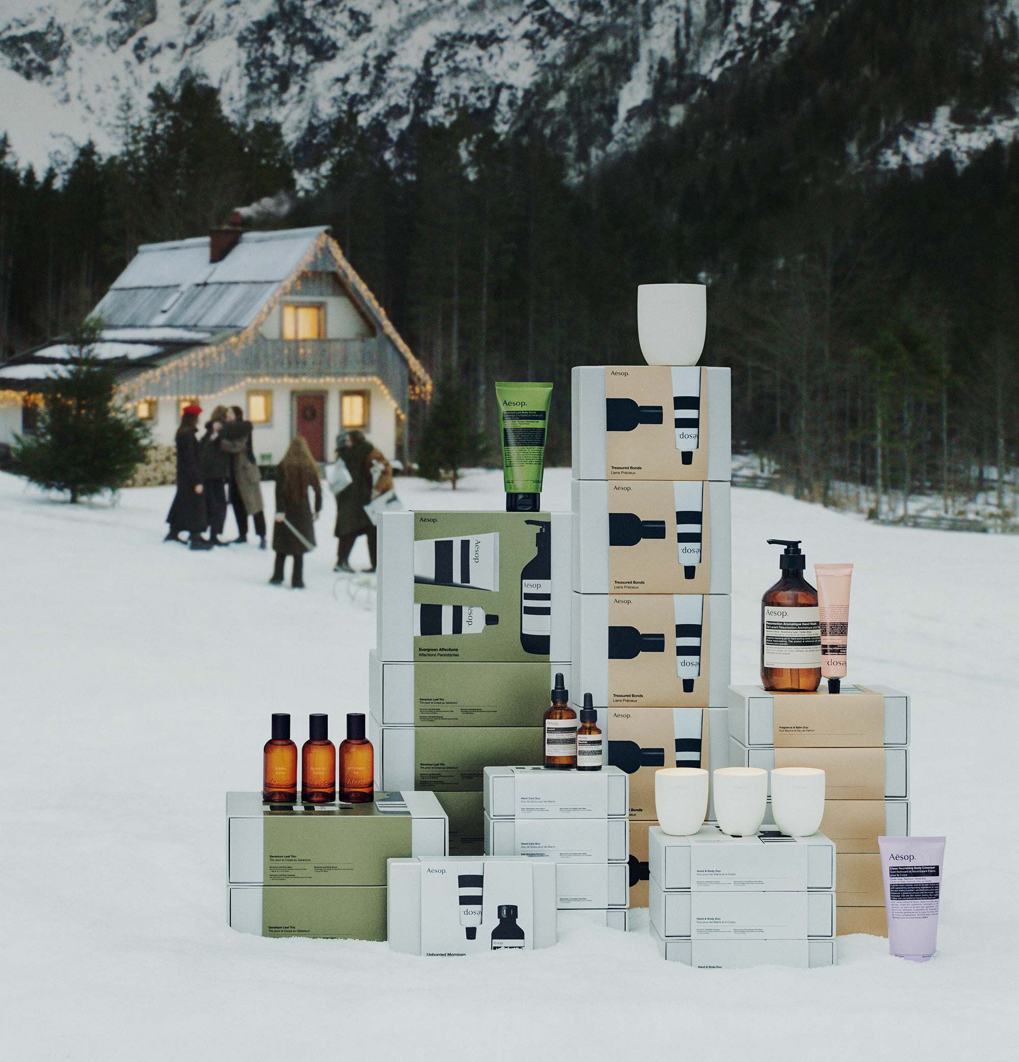 Aesop boxes, bottles, tubes and white candle pots stacked in snow, with people beside a lit house beneath snowy mountains.